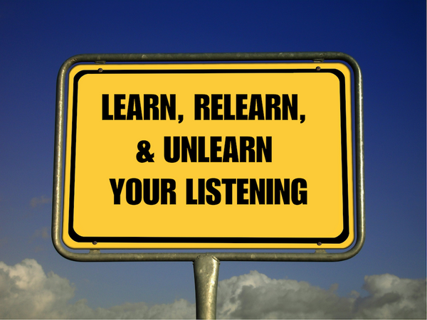 A yellow sign that says, “LEARN, RELEARN, & UNLEARN YOUR LISTENING.” Blue sky is in the background.